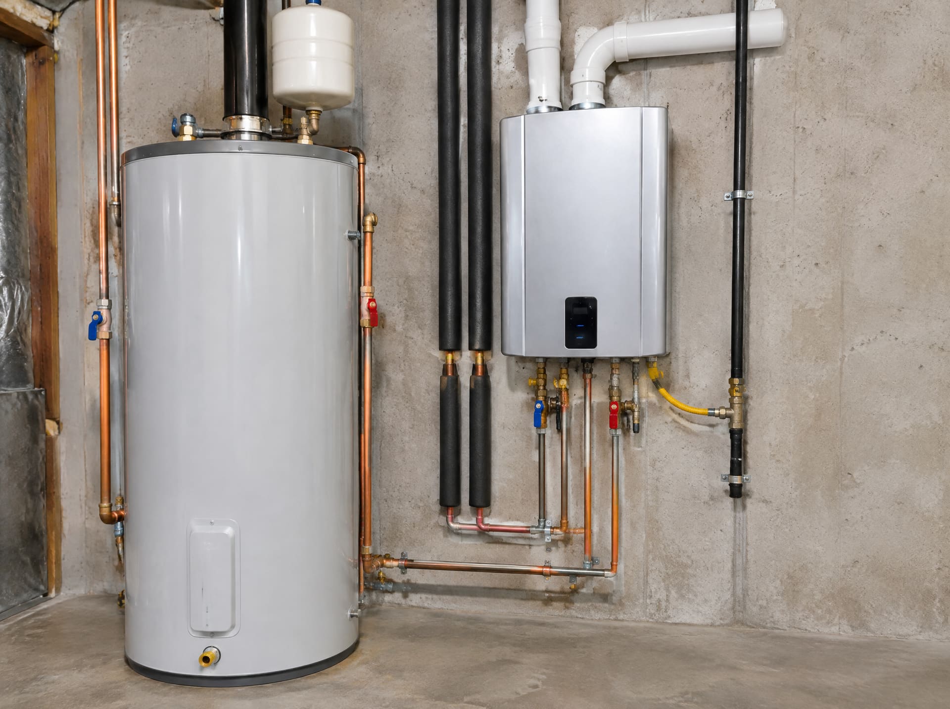 A wall-mounted tankless water heater installed beside a conventional tank in a basement
